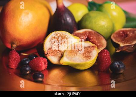 Auf einem Tisch steht ein goldener Teller mit frischen tropischen Früchten. Halbiert geschnittene Feigen, Mango, Limetten und saisonale blaue Beeren, Himbeeren. Vielzahl von s Stockfoto