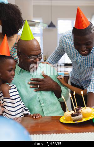 Mehrrassiger älterer Mann mit Partyhut und Blick auf Cupcakes, während er mit der Familie Geburtstag feiert Stockfoto