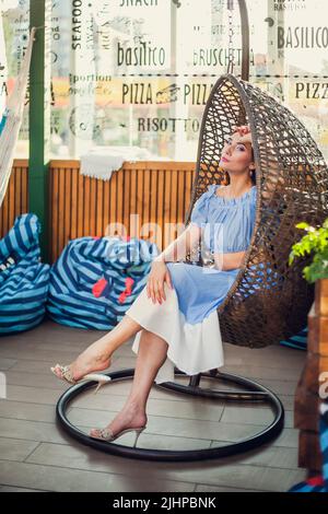 Die wunderschöne junge Frau sitzt in einem Korbstuhl auf der Sommerterrasse eines Stadtcafés, blickt auf die Kamera und lächelt. Stockfoto