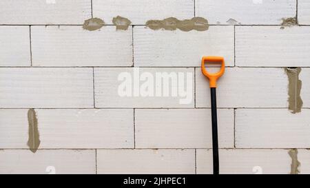 Schwarze Schaufel mit orangefarbenem Griff auf weißem Steinwand-Hintergrund. Spärliche Komposition mit Kopierbereich Stockfoto