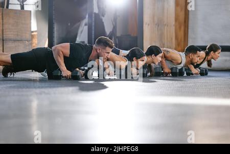 Eine vielfältige Gruppe aktiver junger Menschen, die Push-up-Übungen mit Kurzhanteln machen, während sie gemeinsam in einem Fitnessstudio trainieren. Fokussierte Athleten, die Pressups mit machen Stockfoto