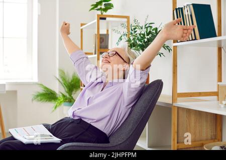Glückliche, entspannte ältere Dame, die auf einem Stuhl sitzt und ihre Arme nach Beendigung der Arbeit streckt Stockfoto