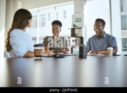 Drei junge, glückliche Geschäftsleute, die bei der Arbeit an einem Tisch sitzen und sich treffen. Fröhliche Geschäftsleute, die im Büro reden und planen Stockfoto