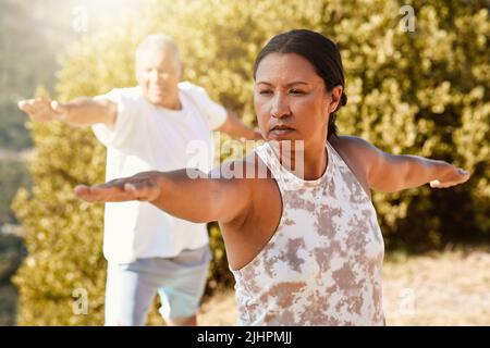 Senioren leben einen gesunden Lebensstil und trainieren im Freien. Das ältere Paar steht in Kriegerpose, während es an einem sonnigen Tag Yoga in der Natur praktiziert Stockfoto