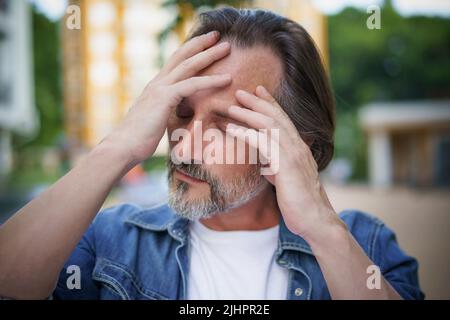 Verzweifelter Mann übergeht die Krise des mittleren Alters und hält seine Stirn emotional erschöpft. Kopfschmerzen, Stress reifer schöner Mann im Freien europäischen alten Stadtstraßen stehen. Stockfoto