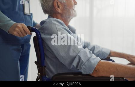 Krankenschwester, die einen Rollstuhl mit einem Fenster für einen älteren Mann aufstellt. Unglücklicher älterer Mann mit geriatrischer oder depressiver Erkrankung. Therapeut kümmert sich, Unterstützung und Stockfoto