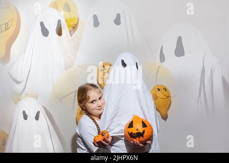 Frohe Halloween. Doppelte Komposition mit Fotos von Mädchen umarmt Geist mit orangenen Kürbis. Kind im weißen Anzug. Lackierte gelbe Kugeln. Emotionen der Freude, des Zorns, des Lachens. Festliches Design, Partykonzept. Stockfoto