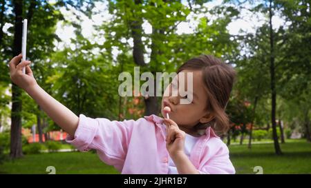 Kind hält Lollipop auf dem Stick, während es Selfie auf dem Smartphone im Park nimmt Stockfoto