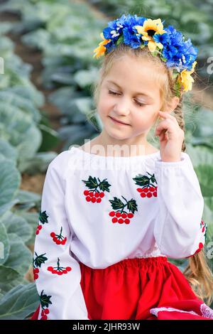 Kindermädchen in der traditionellen ukrainischen Wyschywanka und blau-gelben Kranz von Blumen vor dem Hintergrund des Kohlfeldes. Viburnum auf Kinderkleidung. Unabhängigkeitstag der Ukraine. Landwirtschaft Stockfoto