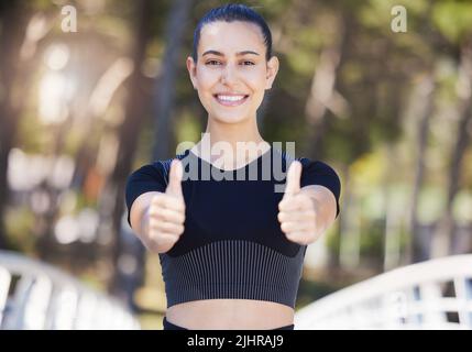 Glückliche weibliche Athletin, die beim Laufen oder Joggen im Freien mit beiden Händen den Daumen nach oben zeigt. Fit junge Frau lächelt und sieht zufrieden während sie Stockfoto