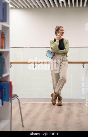 Frau mit Buch in den Händen, die in der modernen öffentlichen Bibliothek auf dem Mobiltelefon spricht Stockfoto