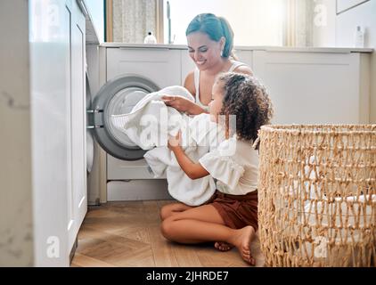 Die junge hispanische Mutter und ihre Tochter sortieren zu Hause schmutzige Wäsche in der Waschmaschine. Bezauberndes kleines Mädchen und ihre Mutter machen gemeinsam Aufgaben Stockfoto