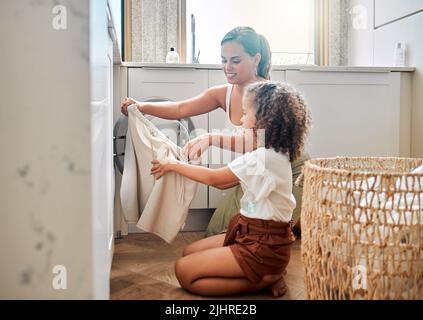 Die junge hispanische Mutter und ihre Tochter sortieren zu Hause schmutzige Wäsche in der Waschmaschine. Bezauberndes kleines Mädchen und ihre Mutter machen gemeinsam Aufgaben Stockfoto