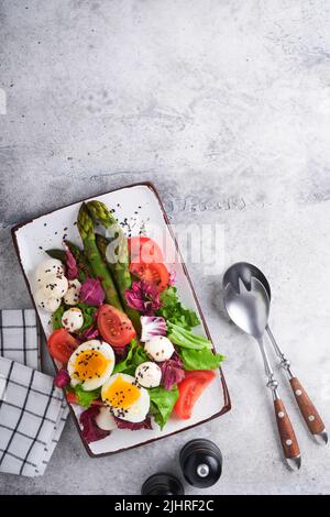 Spargel, Tomaten, Salat, Mozzarella, schwarzer Sesam, Flachs, Ölolivsalat und weich gekochtes Ei auf rechteckiger Keramikplatte auf grauem Betontisch Bac Stockfoto