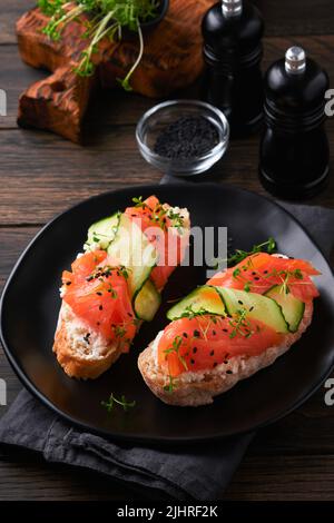 Sandwiches. Lachs-Toast mit Frischkäse, Gurke, schwarzem Sesam und Microgreens auf altem Holztisch-Hintergrund. Meeresfrüchte. Gesunde Ernährung. Fotografie Stockfoto