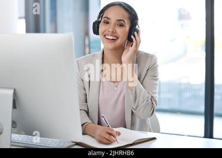 Junge, glückliche Geschäftsfrau mit gemischtem Rennen, die Kopfhörer trägt und Musik hört, während sie allein in einem Büro in einem Notebook schreibt. Ein hispanischer Geschäftsmann Stockfoto