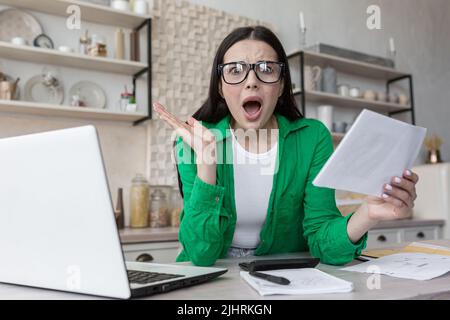 Haushaltsfinanzen, Hausfrau schockiert beim Blick auf die Kamera und schreiend frustriert, bekam einen schlechten Brief von der Bank, Frau hinter Papierarbeit mit Laptop zu Hause in der Küche Stockfoto