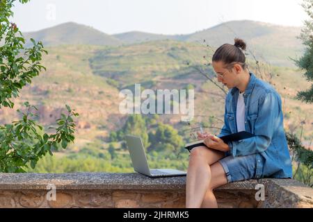 Junger Mann, der als Freiberufler am Computer arbeitet und in seinem Notizbuch zeichnet, umgeben von einer natürlichen Landschaft in europa Stockfoto