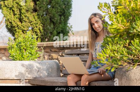 Junge, schöne Frau, die als digitale Nomade auf ihrem Laptop arbeitet, während sie im Sommer auf einer Holzbank im Park sitzt Stockfoto