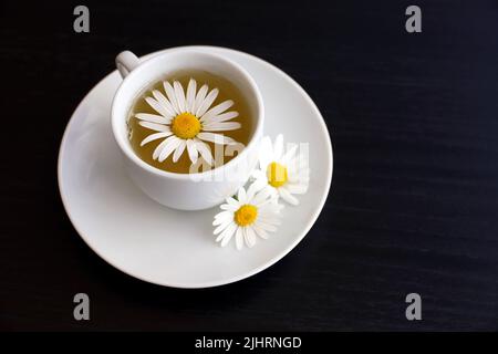 Kamillentee in einer weißen Tasse, Gänseblümchen auf der Untertasse. Heilendes Kräutergetränk auf dunklem Holztisch Stockfoto