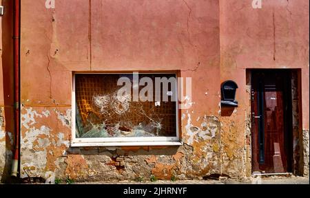 Abblätternde Farbe an einer verfallenen Fassade mit einer Regenrinne, einer Holztür und einem Schaufenster, dessen Glas mehrmals gesprungen ist Stockfoto