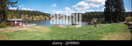 Panoramaaufnahme eines Jagdhauses am Ufer eines kleinen blauen Sees inmitten eines dichten Waldes vor einem klaren, bewölkten Himmel Stockfoto