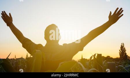 Ein glücklicher und zufriedener junger Bauer auf seinem Maisfeld mit ausgestreckten Händen. Kaukasischer Mann, der auf dem landwirtschaftlichen Feld steht und den Sonnenuntergang genießt Stockfoto