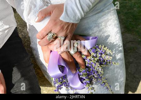 Hände mit Eheringen eines frisch verheirateten Paares mit violettem Strauß Lavendelblüten. Stockfoto