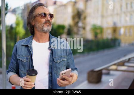 Der Mann mittleren Alters wurde vom Mobiltelefon abgelenkt und genoss Kaffee im Freien und schaute den Sonnenuntergang an. Mann mittleren Alters, der auf die Straße schaut und dabei Freizeit von der Arbeit genießt. Unterwegs. Stockfoto