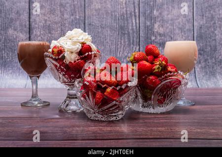 Drei Glasvasen voller reifer roter Erdbeeren sitzen auf einem Holztisch. In der Nähe befinden sich Weingläser mit Milch- und Schokoladencocktails. Stockfoto