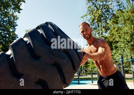 Starker Mann Training Workout Lifting großen Reifen Outdoor-Fitnessraum. Sportlicher junger Erwachsener kaukasischer Kerl Flip großes Rad auf die Natur. Handgefertigte Ausrüstung Sport Stockfoto