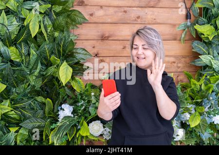 Eine lächelnde asiatische Frau in einer schwarzen Jacke nutzt ein Smartphone für die Online-Videokommunikation und winkt mit der Hand, um Freunde zu grüßen. Online-Kommunikation mit Stockfoto