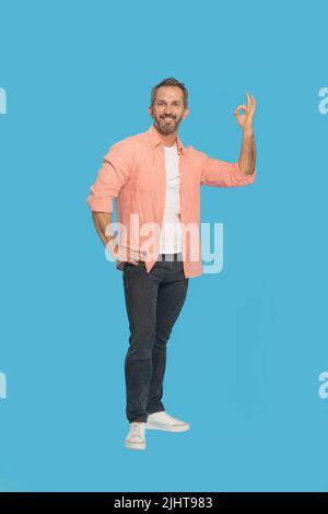 Gestikulieren OK schöner Mann im mittleren Alter grauhaarig glücklich lächelnd Blick auf Kamera trägt Pfirsich Hemd isoliert auf blauem Hintergrund. Fit reifen Mann sagen im OK. Stockfoto