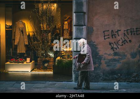 Eine alte Dame, die an einem schicken Modeladen vorbeiläuft und an der angrenzenden Wand Graffiti mit der Aufschrift „Eat the Rich“ aufführt Stockfoto