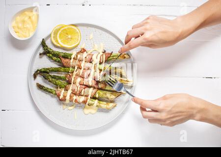 Gegrillter grüner Spargel, eingewickelt mit Speck. Weibliche Hände halten Messer und Gabel. Ketogene Ernährung. Gesunde Ernährung, Diät-Menü. Draufsicht, Overhead Stockfoto