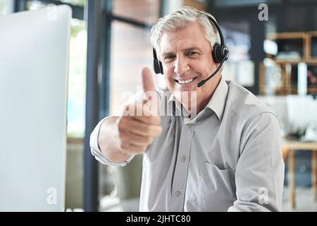 Jahrelange Erfahrung am Arbeitsplatz. Ein ausgereifter Call Center-Agent sitzt allein im Büro und zeigt den Daumen nach oben. Stockfoto
