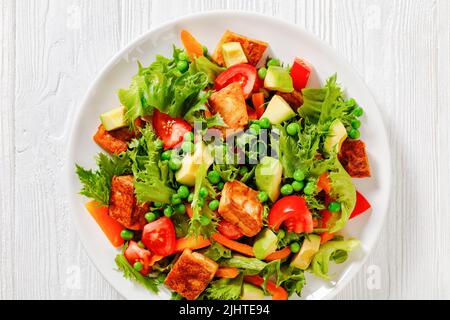 Tofu-Salat mit Avocado, knackigen Salatblättern, grünen Erbsen, Tomaten, gelber Paprika auf weißem Teller, flach liegend, Nahaufnahme Stockfoto