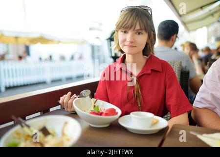 Hübsches Teenager-Mädchen, das am sonnigen Sommertag im Freien leckeres frisches Eis isst. Köstliches und appetitliches Eis-Dessert auf einem Tisch. Kaltes Karamelleis Stockfoto