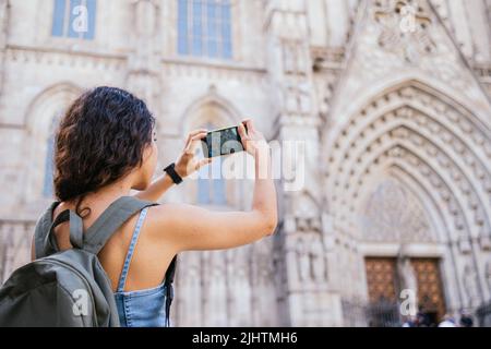 Junge Brünette mit Rucksack fotografieren mittelalterliche Kathedrale per Smartphone, während die Straßen der historischen Stadt tagsüber erkunden Stockfoto
