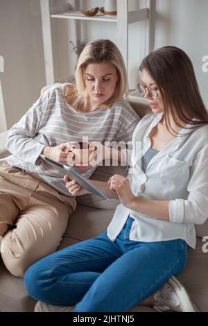 Zwei junge Freundinnen, die zusammen ein Tablet benutzen, sitzen auf dem Co Stockfoto