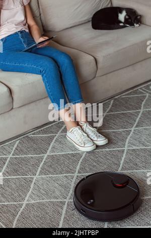 Roboter-Staubsauger Reinigung Teppich, Frau mit Tablet und Freude Rest und Katze sitzt auf dem Sofa zu Hause Stockfoto