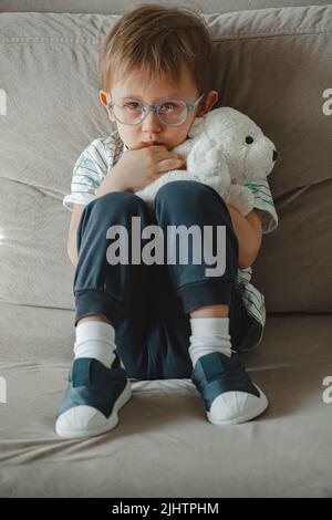 Ein Kind mit Autismus in einer Brille sitzt auf dem Sofa und ist traurig, umarmt sich Stockfoto