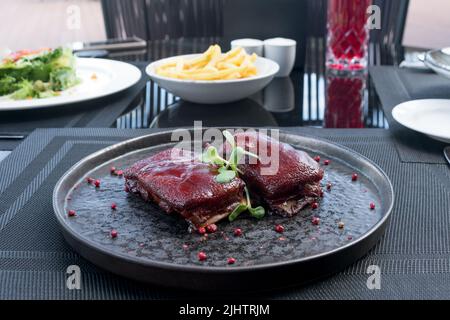 Gegrillte Schweineribs mit süßer und saurer karamellisierter gegrillter Sauce, Rauch, Gewürzen und Mikrogemüse. Grillrippen auf einem schwarzen Teller. Amerikanisches Essen. Stockfoto