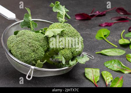 Brokkoli-Kohl im Sieb. Spinatblätter auf dem Tisch. Oberkante Schwarzer Hintergrund Stockfoto