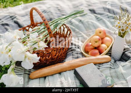 Picknickkorb aus Korb mit Essen und grauer Decke auf Gras im Park. Picknick-Konzept. Vorderansicht. Horizontale Zusammensetzung. Stockfoto