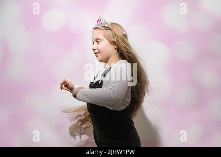 Happy Birthday girl mit langen blonden fließenden Haaren und Krone auf dem Kopf dreht sich mit Freude vor dem Hintergrund der hellen rosa Wand. Das Kind feiert Weihnachten oder den Muttertag. Weicher, selektiver Fokus. Speicherplatz kopieren Stockfoto