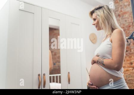 Glückliche Schwangerschaft. Kaukasische blonde Millennial bald-zu-werden Mutter vor einem Kleiderschrank Spiegel stehen und Blick auf ihren großen Schwangerschaftsbauch. Hochwertige Fotos Stockfoto