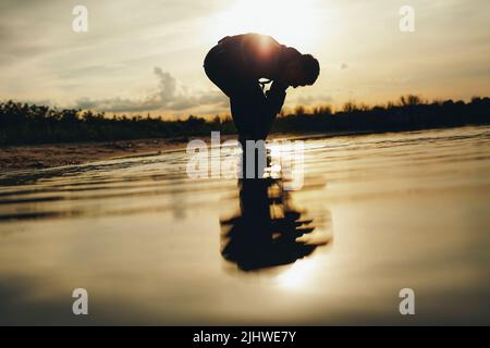 Männlicher Taucher, der am Strand im Wasser steht und Tauchflossen aufsetzt. Abenteuerlicher junger Mann, der sich auf einen Tauchgang bei Sonnenuntergang vorbereitet. Stockfoto