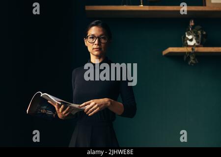 Selbstbewusste junge Frau, die ein Magazin hält und in die Kamera blickt. Geschäftsfrau steht mit Buch im Büro. Stockfoto
