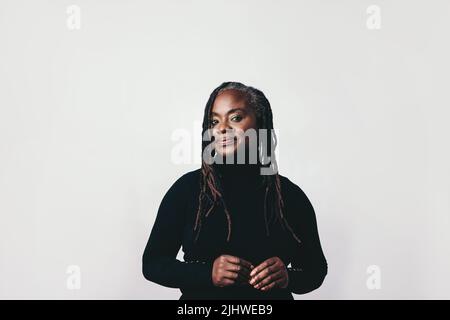 Selbstbewusste Frau mittleren Alters mit Dreadlocks, die in einem Studio die Kamera anschaut. Elegante schwarze Frau, die vor einem grauen Hintergrund steht. Stockfoto
